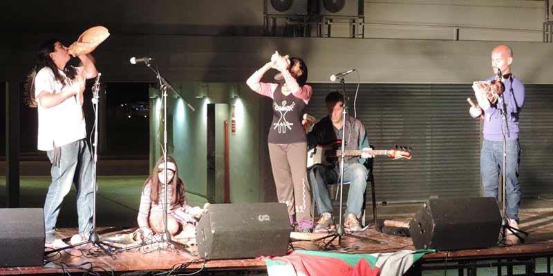 Ciro y el Clan Choñik en un escenario. A la izquierda, Ciro tocando un instrumento musical de viento tradicional, la caparzón de un caracol de mar, en el centro, sentada, una niña indígena tocando instrumentos de percusión, a la derecha, una compañera artista de Ciro tocando también un instrumento de viento, y a la derecha de ella, otro artista tocando un instrumento de percusión, al fondo acompaña un músico tocando el bajo sentado.